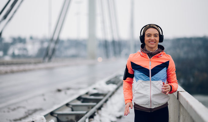 Jeune femme souriante en jogging, un casque sur les oreilles, qui cour sur un pont suspendu enneigé.