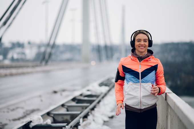 Jeune femme souriante en jogging, un casque sur les oreilles, qui cour sur un pont suspendu enneigé.